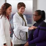 Two women in white coats are using a stethoscope to examine a patient